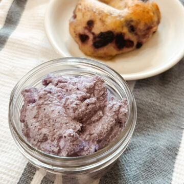 Homemade Raw Milk Blueberry Cream Cheese in a small mason jar next to a homemade sourdough blueberry bagel