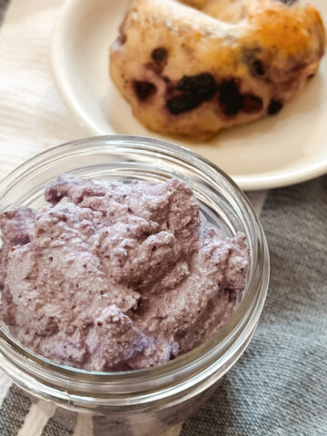 Homemade Raw Milk Blueberry Cream Cheese in a small mason jar next to a homemade sourdough blueberry bagel